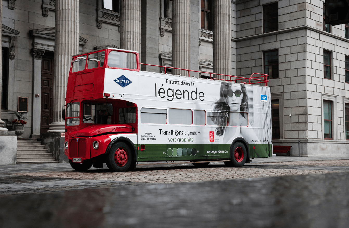 Autobus rouge habillé d'une publicité Transitions Signature Entrez dans la légende avec un grand visuel d'une jeune fille portant des verres les cheveux dans le vent.