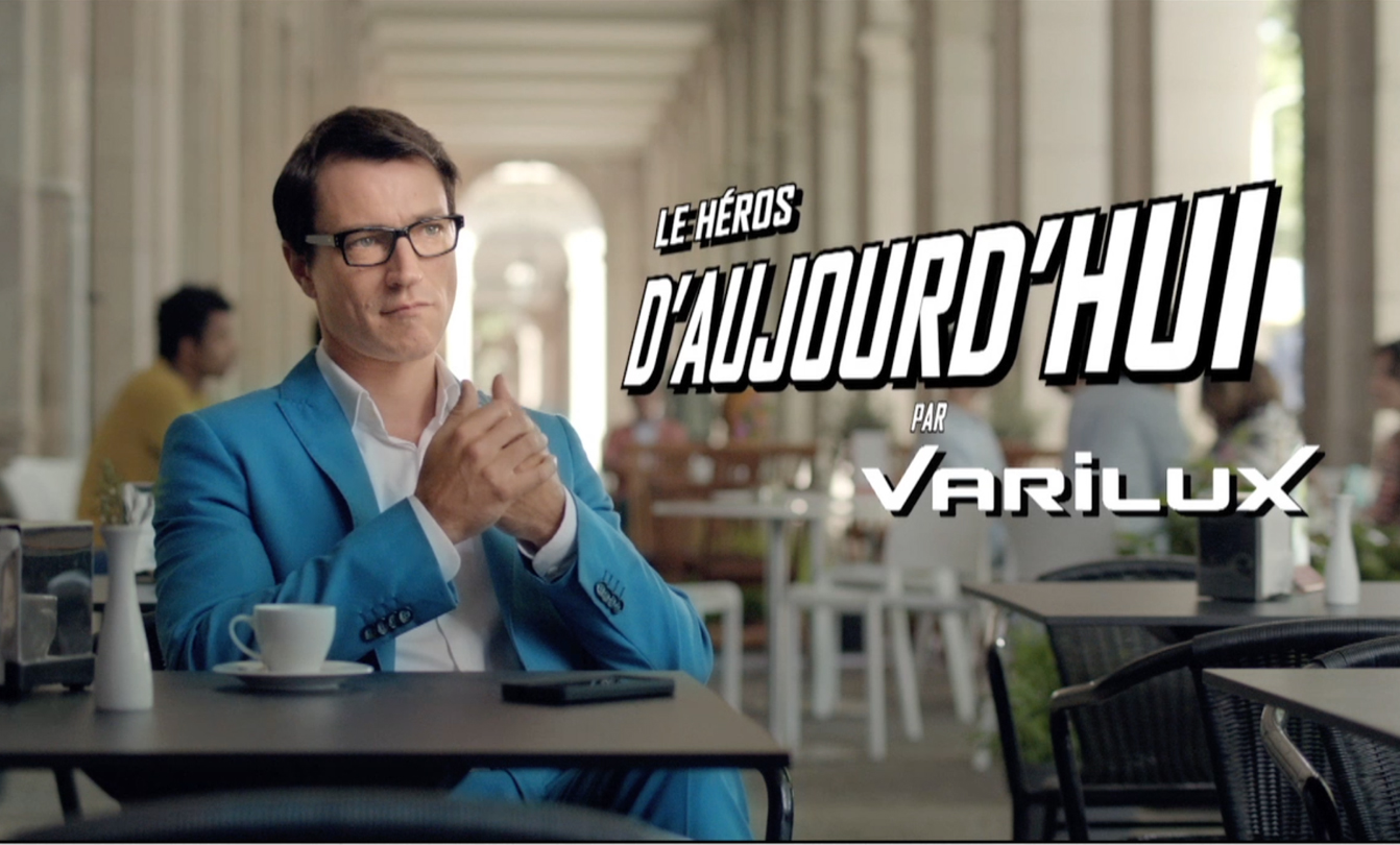 Un homme au veston bleu ciel assis à un café avec slogan Les héros d'aujourd'hui par Varilux